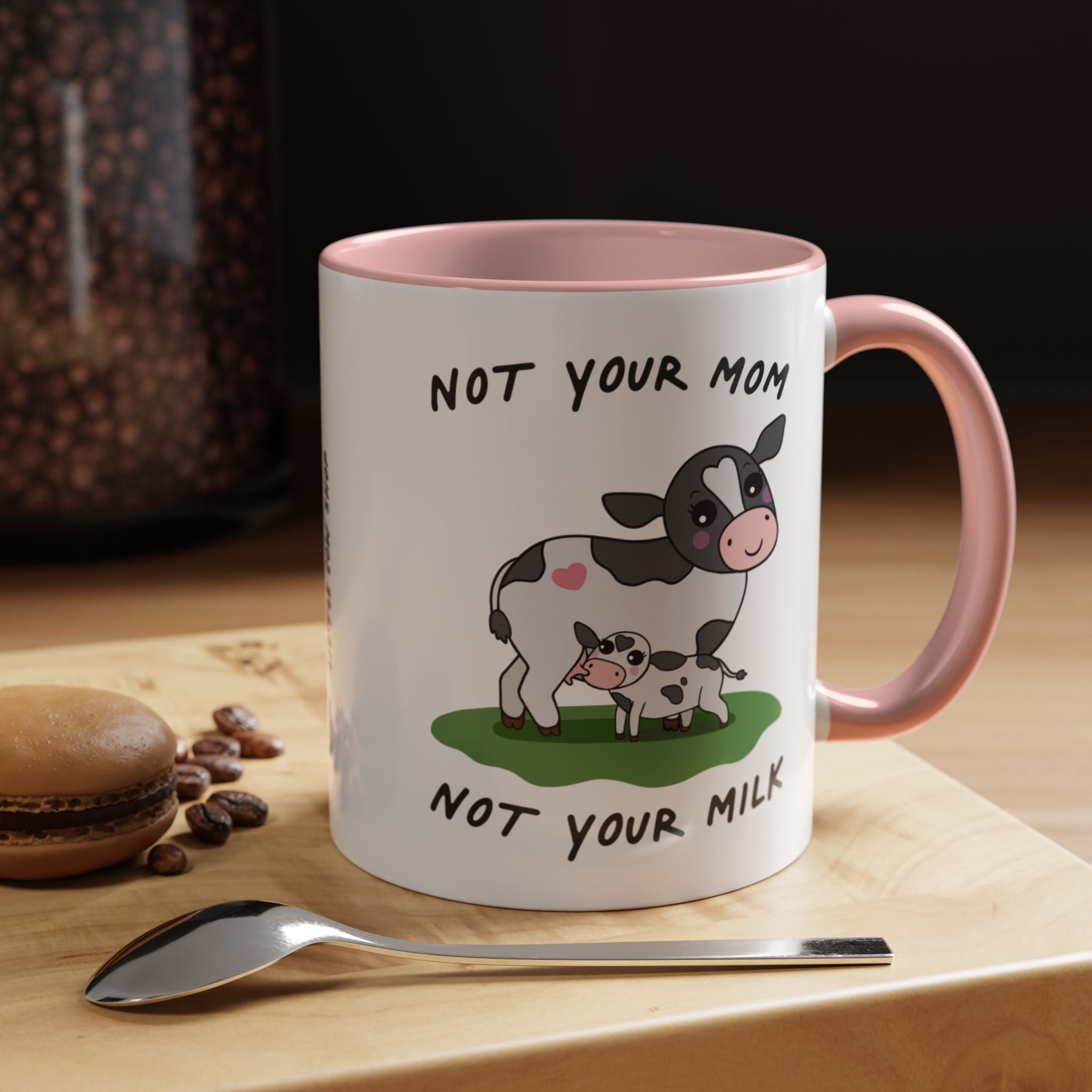 A white ceramic coffee mug with a pink handle and interior, featuring a cartoon cow and calf illustration with the text 'NOT YOUR MOM / NOT YOUR MILK'. The mug sits on a wooden board with a spoon, coffee beans, and a macaron in a warm, styled setting.