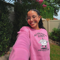 A woman smiling for a selfie in a backyard, wearing a bright pink sweatshirt with a graphic of a cake and the text 'ALL YOU NEED IS LOVE AND VEGAN CAKE'.