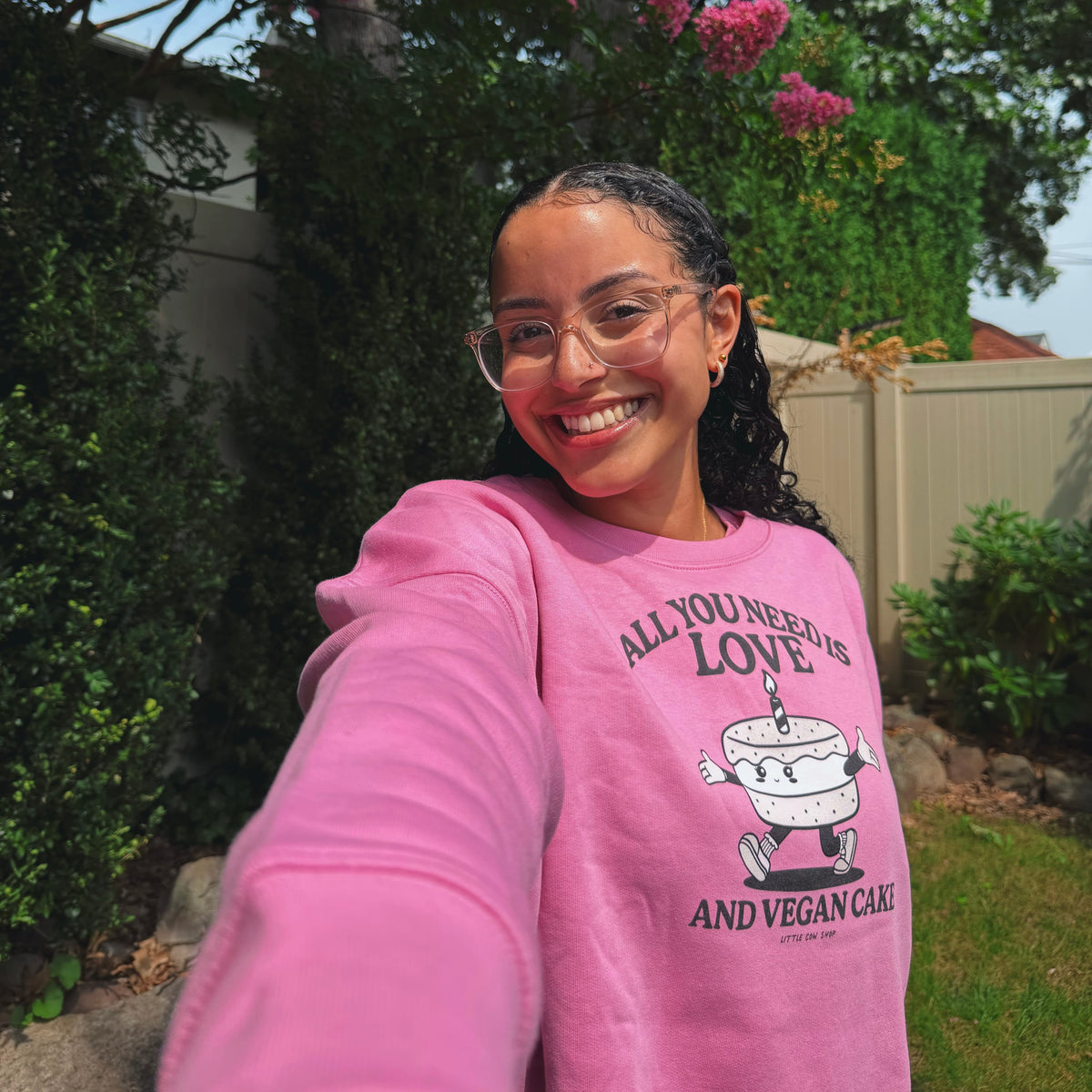 A woman smiling for a selfie in a backyard, wearing a bright pink sweatshirt with a graphic of a cake and the text 'ALL YOU NEED IS LOVE AND VEGAN CAKE'.