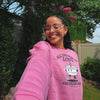 A woman taking a selfie outdoors wearing a bright pink crewneck sweatshirt with a cartoon cake graphic and the text 'ALL YOU NEED IS LOVE AND VEGAN CAKE'.