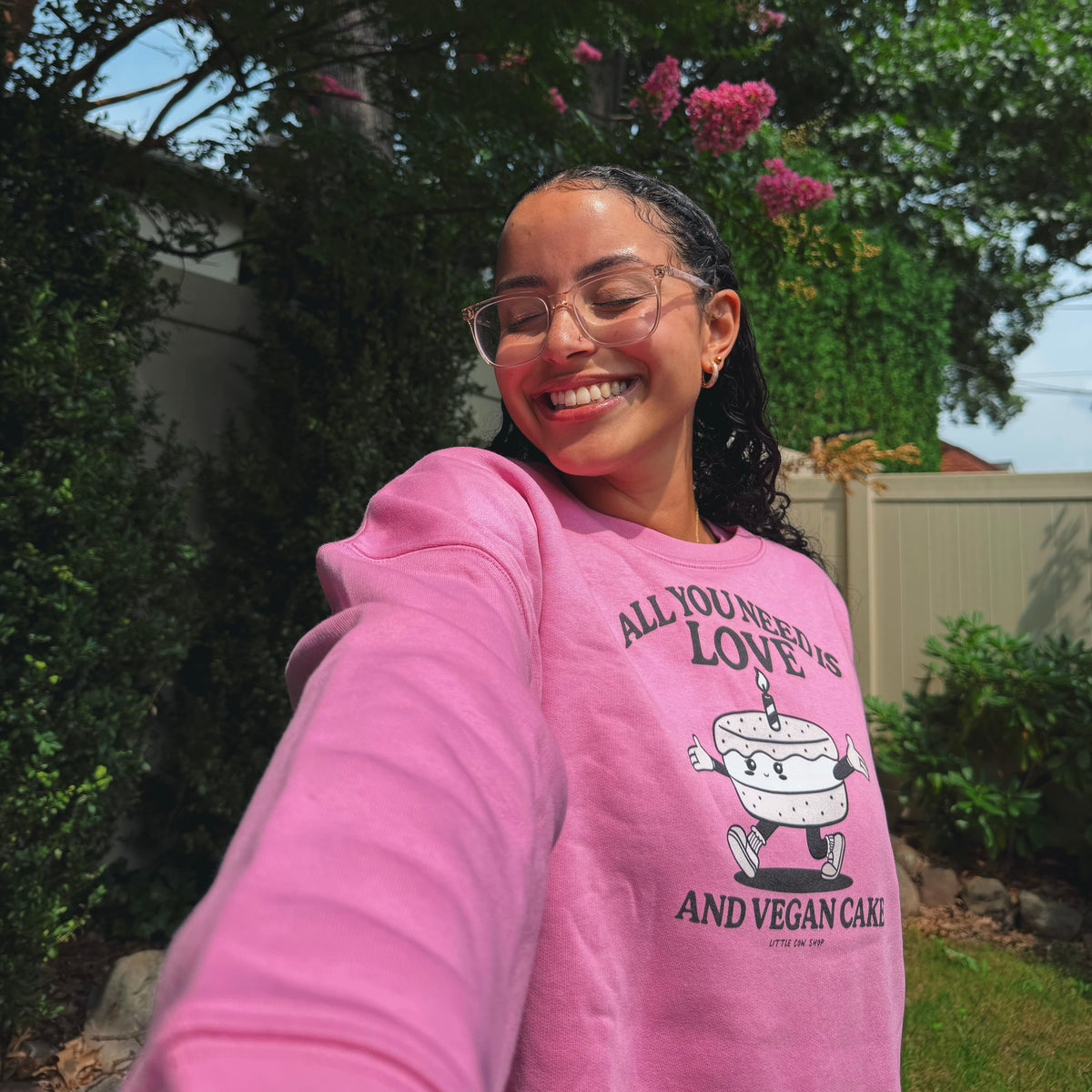 A woman taking a selfie outdoors wearing a bright pink crewneck sweatshirt with a cartoon cake graphic and the text 'ALL YOU NEED IS LOVE AND VEGAN CAKE'.
