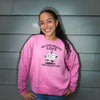 A smiling young woman with glasses stands against a gray siding wall wearing a pink crewneck sweatshirt with a cartoon cake graphic and the text 'ALL YOU NEED IS LOVE AND VEGAN CAKE'.