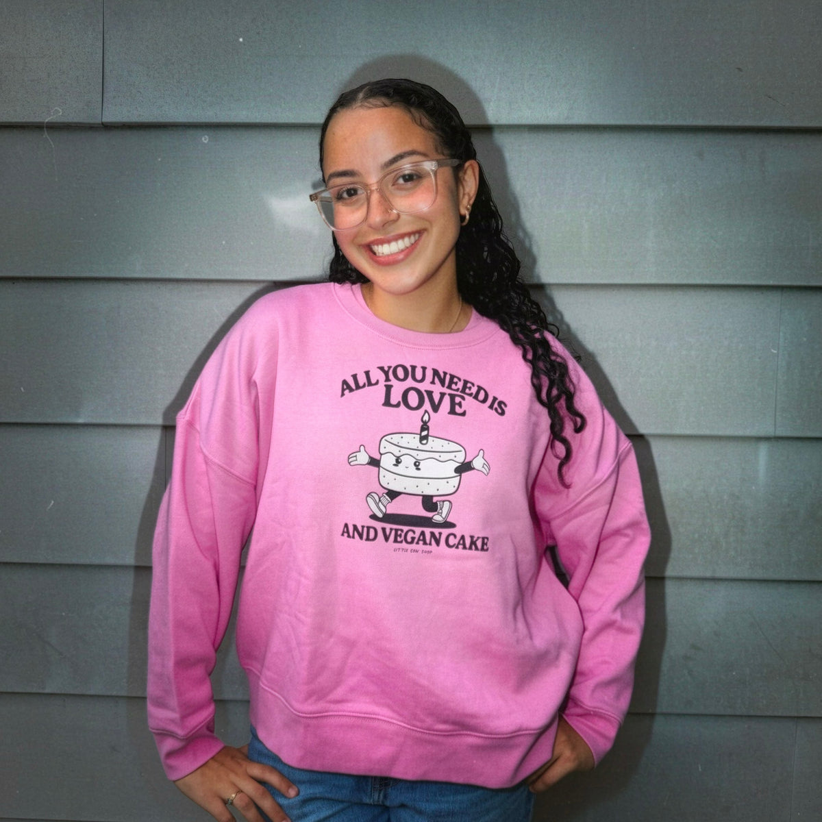 A smiling young woman with glasses stands against a gray siding wall wearing a pink crewneck sweatshirt with a cartoon cake graphic and the text 'ALL YOU NEED IS LOVE AND VEGAN CAKE'.