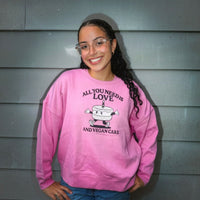 A smiling young woman with glasses stands against a gray siding wall wearing a pink crewneck sweatshirt with a cartoon cake graphic and the text 'ALL YOU NEED IS LOVE AND VEGAN CAKE'.