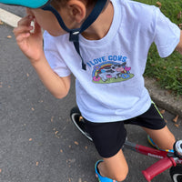 A child wearing a white graphic T-shirt with a colorful cartoon cow and rainbow design that reads 'I LOVE COWS', paired with black shorts and a teal helmet, riding a red balance bike on an outdoor paved path.