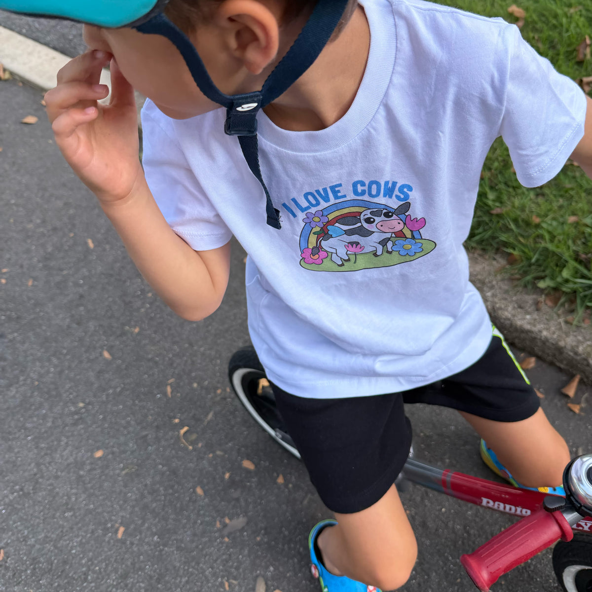 A child wearing a white graphic T-shirt with a colorful cartoon cow and rainbow design that reads 'I LOVE COWS', paired with black shorts and a teal helmet, riding a red balance bike on an outdoor paved path.