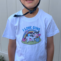 A child wearing a white short-sleeve graphic T-shirt with a colorful cartoon cow, rainbow, flowers, and the text 'I LOVE COWS' on the front, photographed against a light paneled wall.