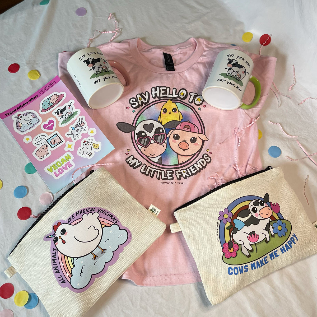 A styled flat-lay of a pink cartoon t-shirt and matching printed accessories on a white fabric background with confetti. The set includes a pink t-shirt reading 'Say Hello to My Little Friends,' two white mugs with cow illustrations, a sticker sheet, and two canvas pouches with cow and unicorn graphics.