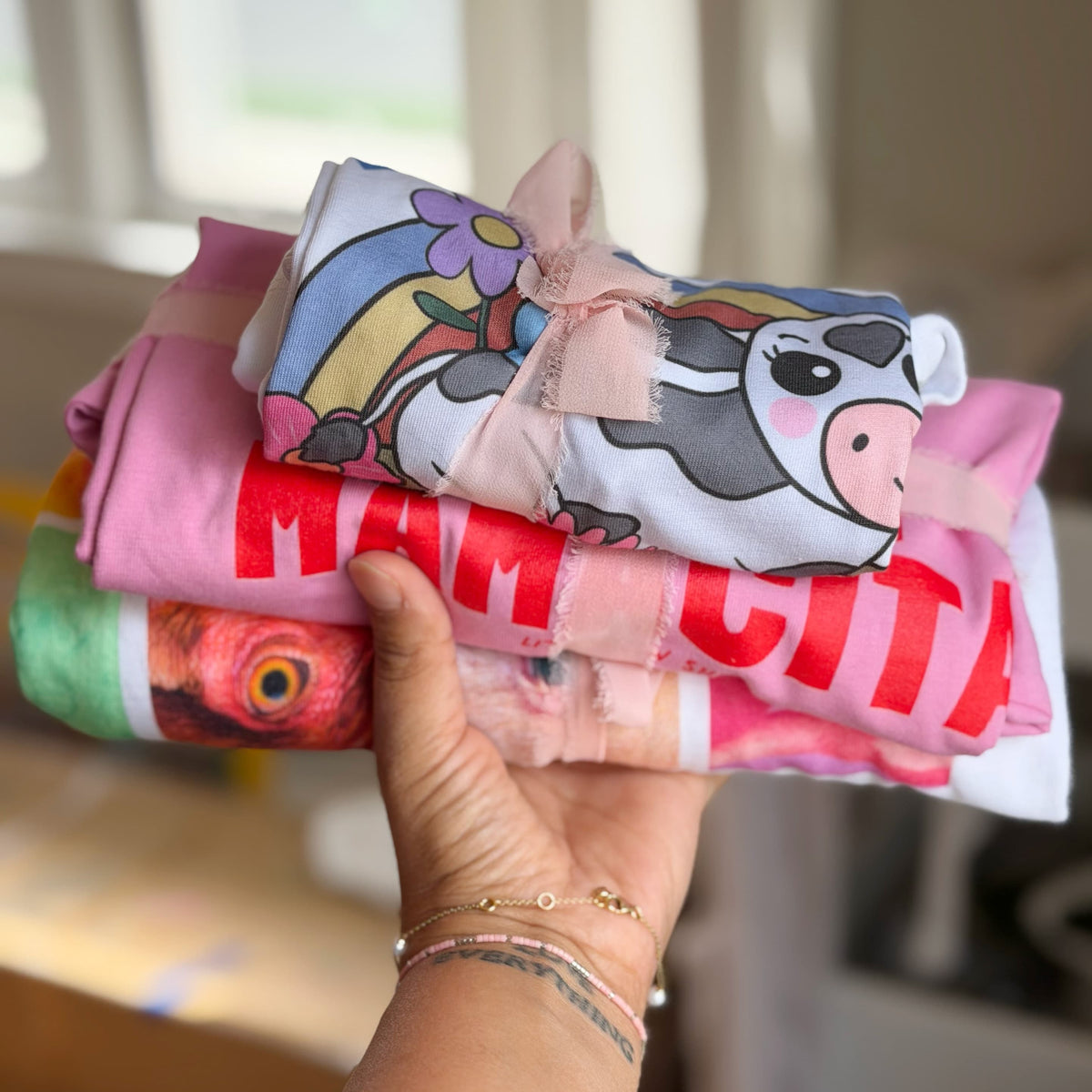 A hand holds a stack of folded colorful garments tied with a sheer pink ribbon. The top garment features a cartoon cow graphic, and the stack shows other bright colors and text designs, photographed in a softly blurred indoor setting.