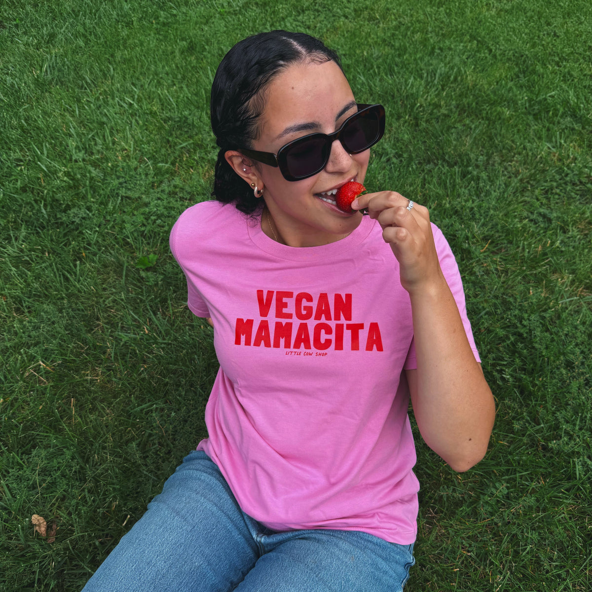 A woman wearing a bright pink graphic t-shirt with the text 'VEGAN HAMACITA' in red, sitting on a green grass lawn and eating a strawberry. She is also wearing black sunglasses and blue jeans.
