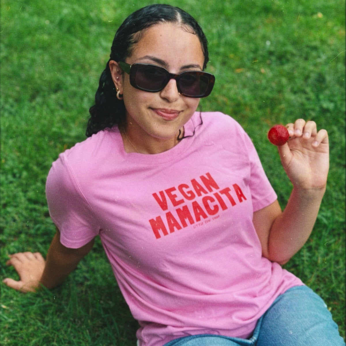 A woman wearing a pink graphic T-shirt with 'VEGAN MAHACITA' printed in red, sitting on green grass and holding a strawberry.