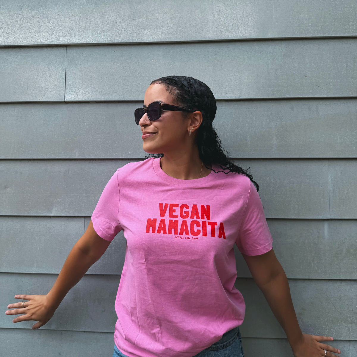 A woman wearing a pink graphic t-shirt with 'VEGAN NAMACITA' printed in red across the chest, posing against a gray horizontal siding wall.