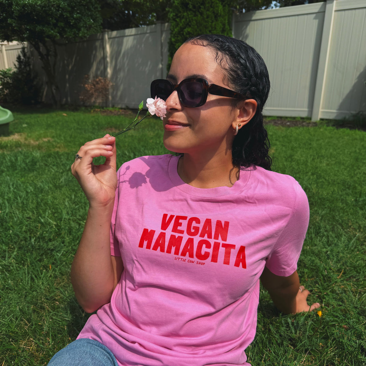 A woman sitting on a green lawn wearing a pink t-shirt with 'VEGAN MAMACITA' printed in red text, holding a small pink flower near her face.