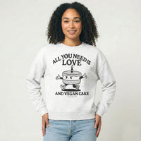 Person wearing a gray sweatshirt with a graphic of a cake character and the text "All you need is Love and vegan cake" on a white background 