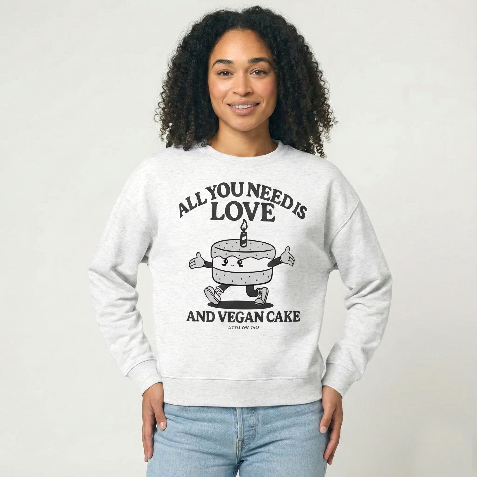 Person wearing a gray sweatshirt with a graphic of a cake character and the text "All you need is Love and vegan cake" on a white background 
