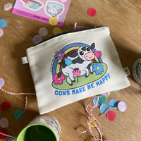 A cream-colored canvas pouch with a cartoon cow, rainbow, and flowers printed on the front with the words "Cows Make me Happy" underneath,  sitting on a wooden table surrounded by colorful confetti and party decorations.