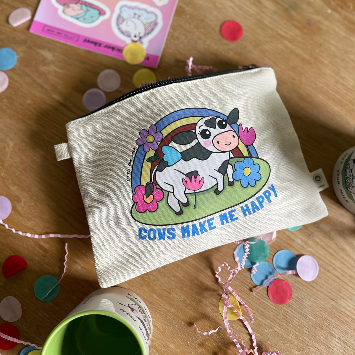 A cream-colored canvas pouch with a cartoon cow, rainbow, and flowers printed on the front with the words "Cows Make me Happy" underneath,  sitting on a wooden table surrounded by colorful confetti and party decorations.