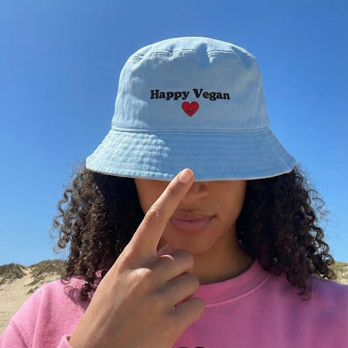 Person wearing a light blue bucket hat with 'Happy Vegan' text and a red heart on a clear blue sky background.