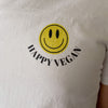 A close-up of a white t-shirt with a graphic design featuring a bright yellow smiley face and the words "HAPPY VEGAN" printed below it in black.