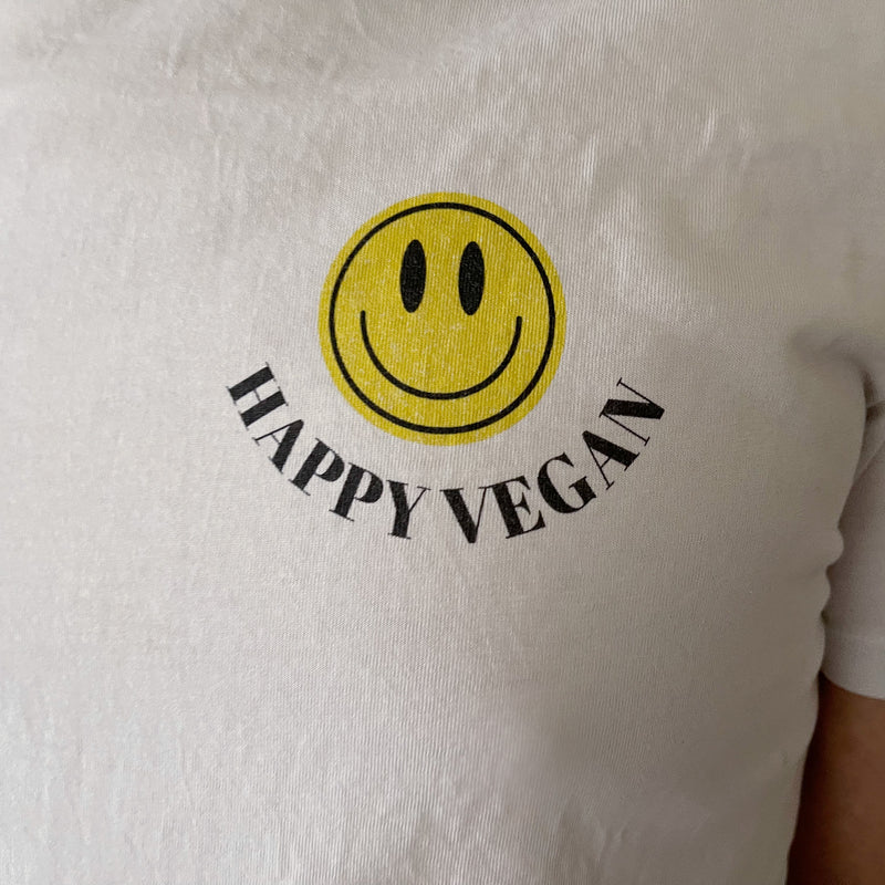 A close-up of a white t-shirt with a graphic design featuring a bright yellow smiley face and the words "HAPPY VEGAN" printed below it in black.