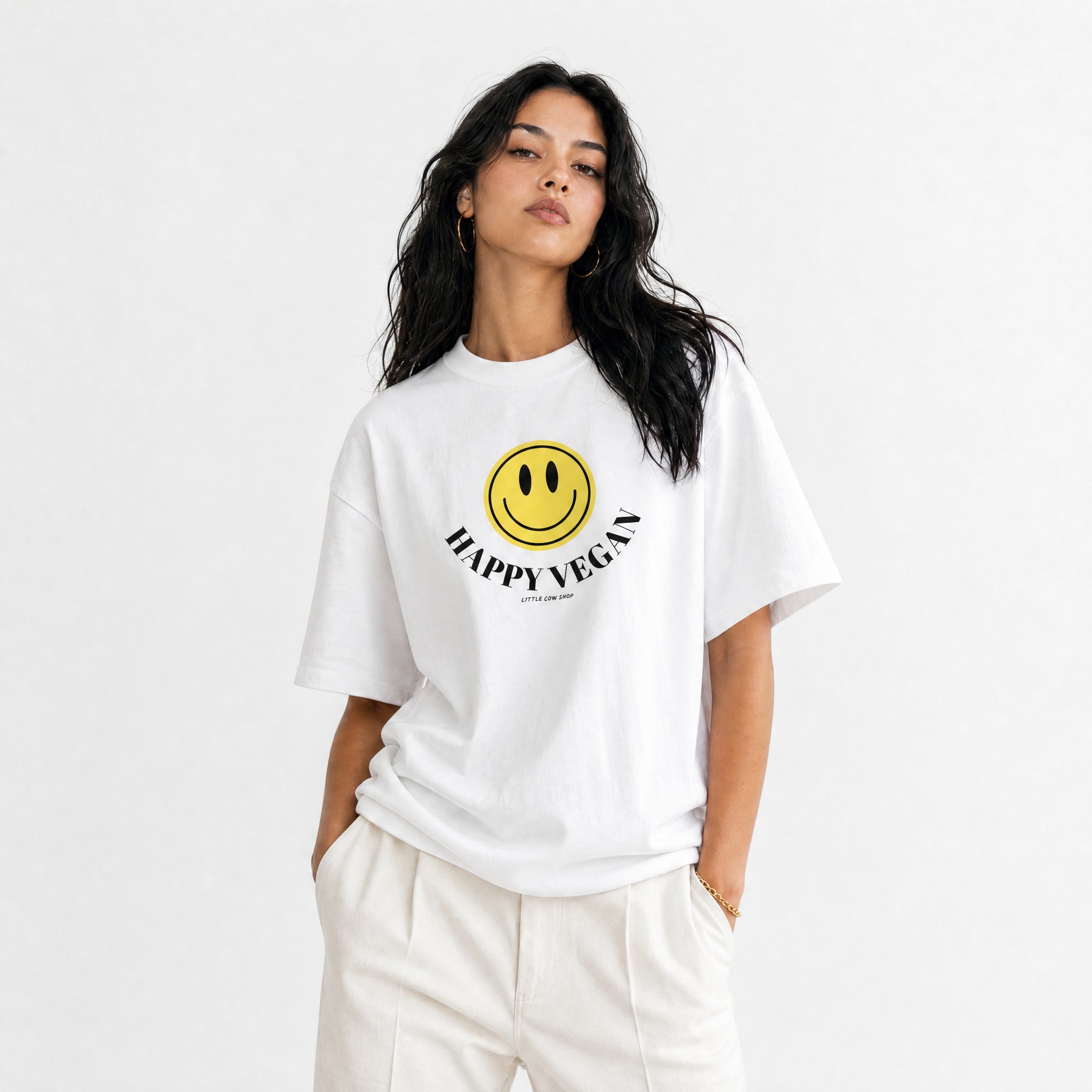 A woman wearing a white oversized T-shirt with a yellow smiley face graphic and the text 'HAPPY VEGAN' printed on the front, paired with white pants, against a plain light gray background.