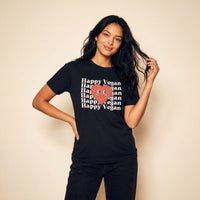 A woman modeling a black graphic t-shirt with 'Happy Vegan' printed in white text surrounding a smiley red heart, set against a plain beige background.