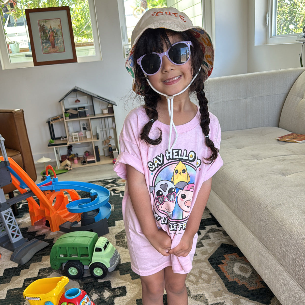 Child in a pink t-shirt with Bo and Friends illustration in the front, with the words "say hello to my little friends", sunglasses, and a hat in a living room with toys.