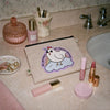 A beige canvas cosmetic pouch with a cartoon unicorn graphic reading 'ALL ANIMALS ARE MAGICAL UNICORNS' is placed on a bathroom counter alongside lipsticks, a compact, and other toiletries.