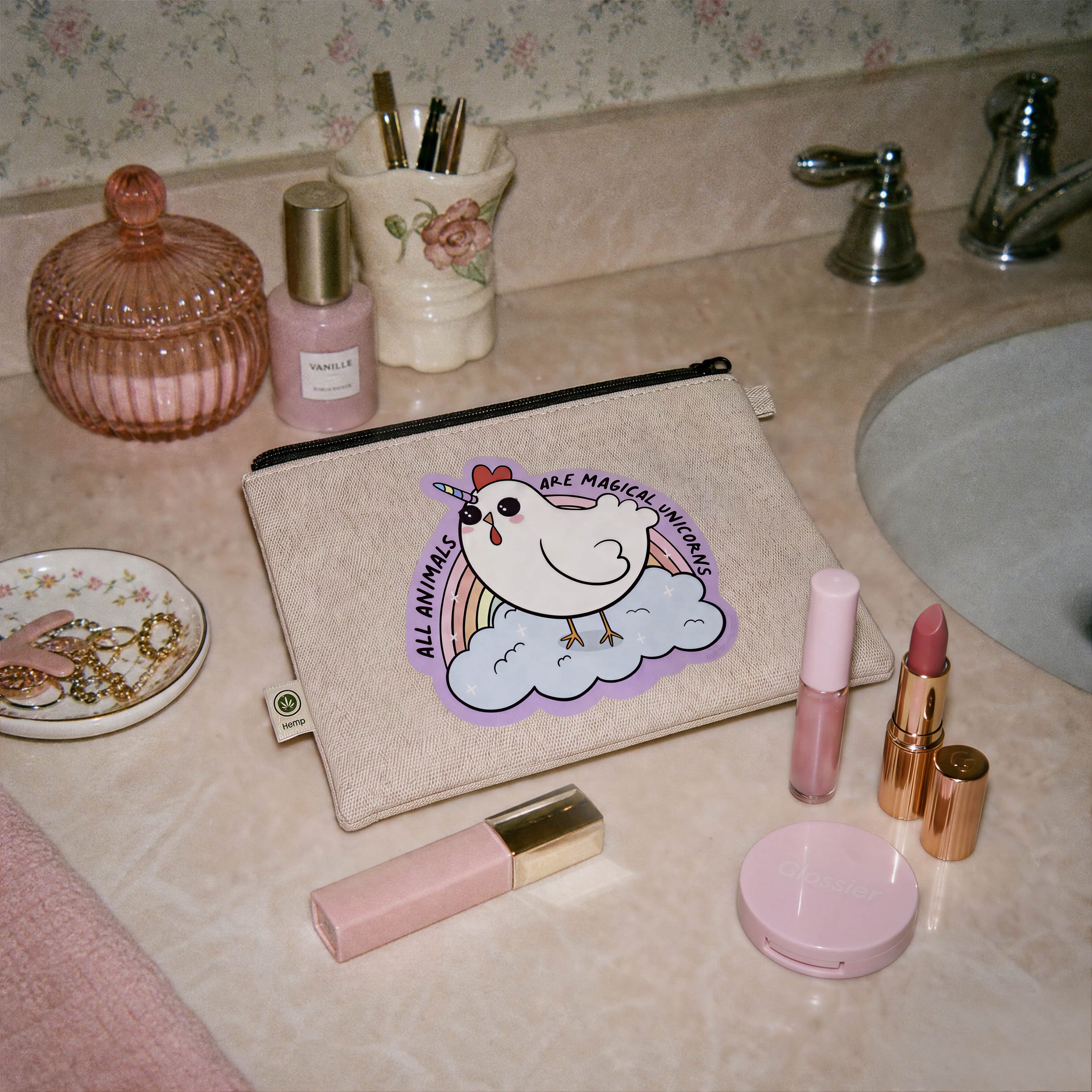 A beige canvas cosmetic pouch with a cartoon unicorn graphic reading 'ALL ANIMALS ARE MAGICAL UNICORNS' is placed on a bathroom counter alongside lipsticks, a compact, and other toiletries.