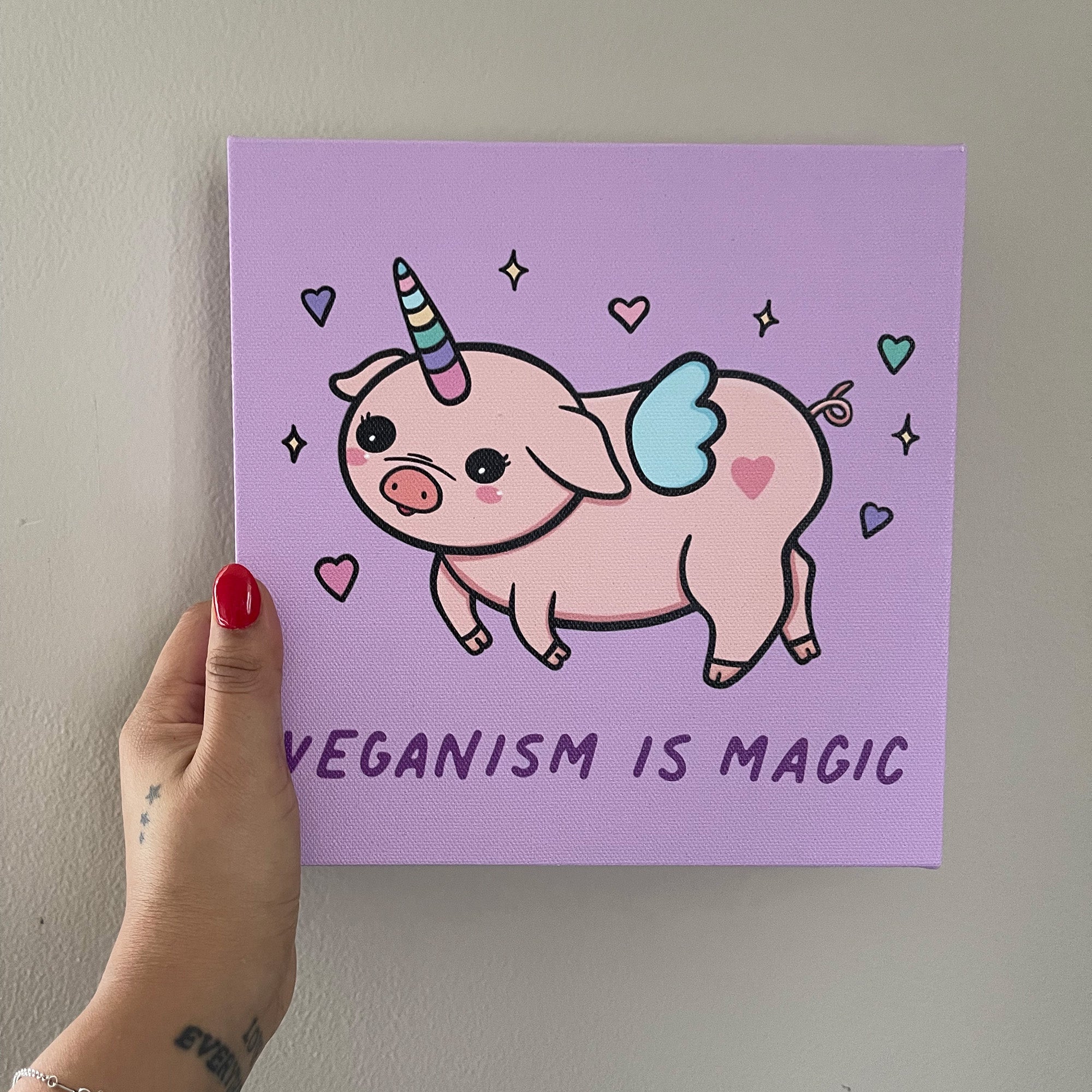 Whimsical illustration of a pink piglet with a rainbow unicorn horn and tiny wings, surrounded by hearts and stars. The background is light purple and the text at the bottom reads 'Veganism is Magic' in a playful font.