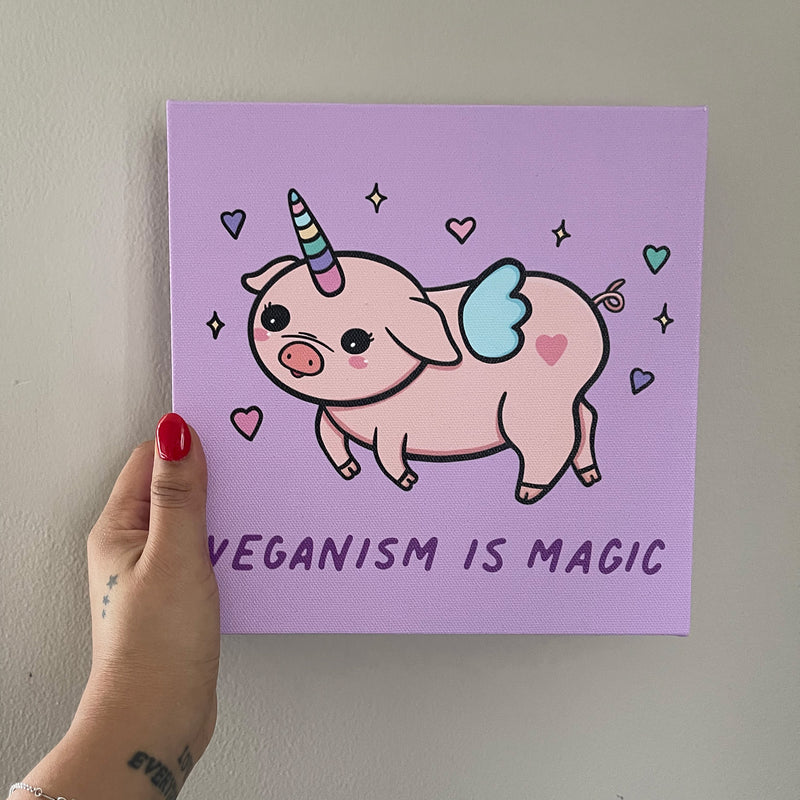 Whimsical illustration of a pink piglet with a rainbow unicorn horn and tiny wings, surrounded by hearts and stars. The background is light purple and the text at the bottom reads 'Veganism is Magic' in a playful font.